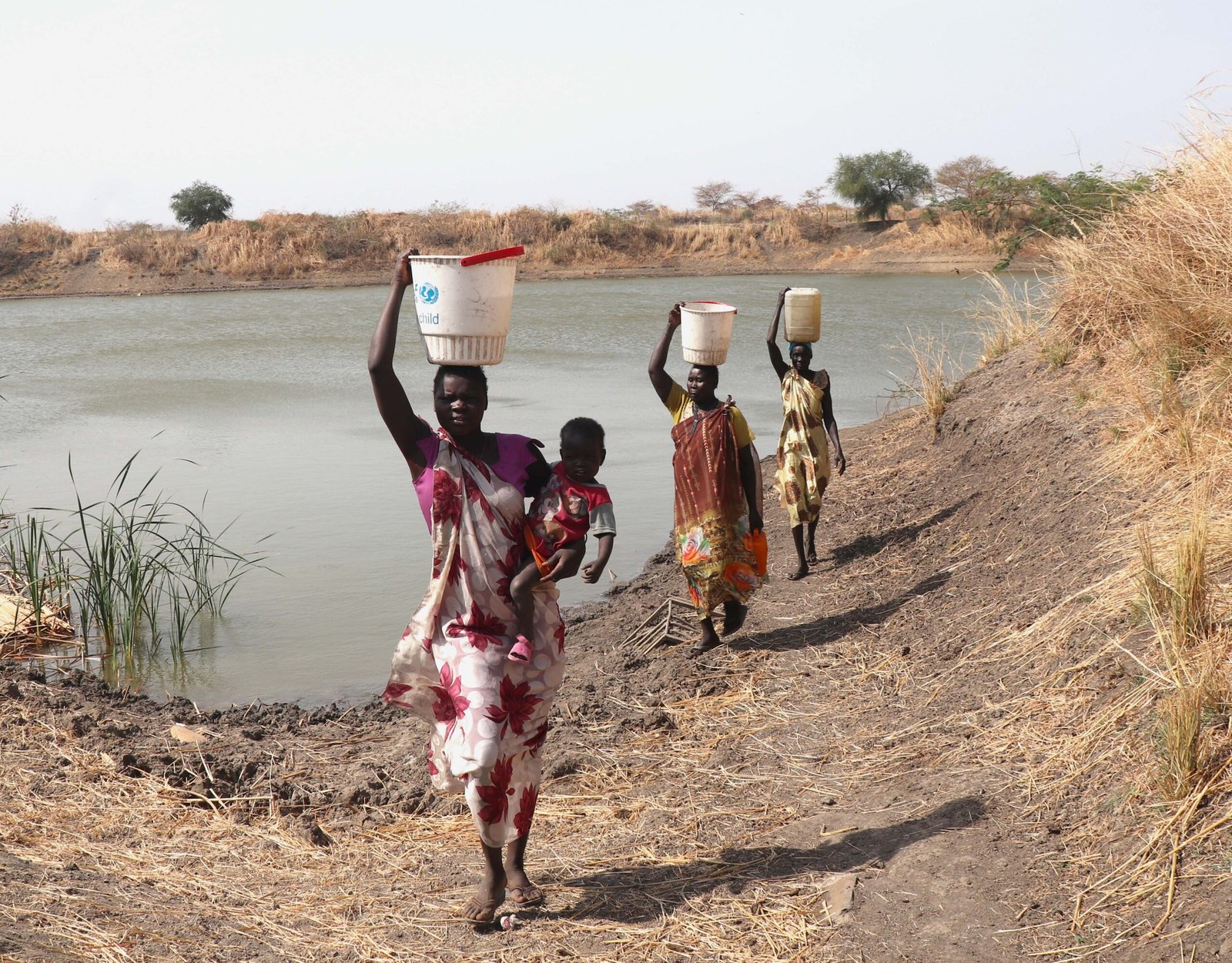 A Sudanese mother and her children walking across cracked sun-baked earth carrying water containers, Darfur, 2026